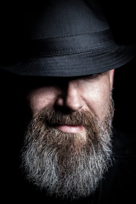 Bart Mann Mann mit Schwarz-Hut und grauem Bart, lächelt geheimnisvoll in dunklem Licht.