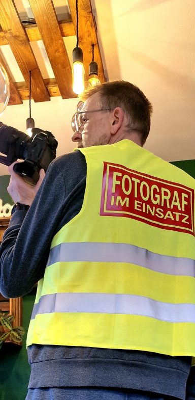 Ein Mann in einem gelben Warnweste mit der Aufschrift "FOTOGRAF" hält eine Kamera.