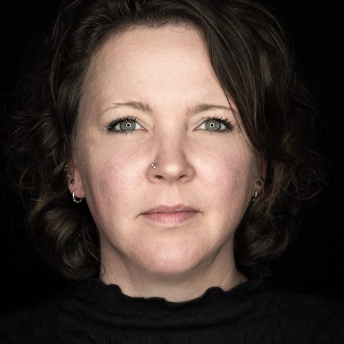 Kundenstimmen Nina Porträt einer Frau mit kurzem, lockigem Haar und neutralem Ausdruck auf schwarzem Hintergrund.