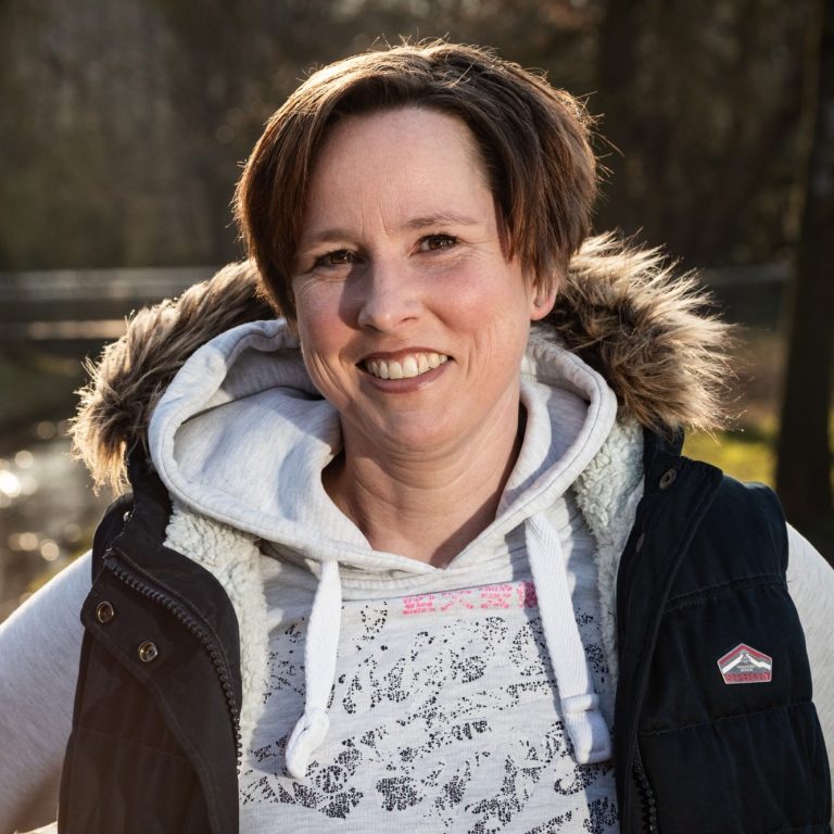 Kunde Tanja Frau mit lässiger Kleidung und Pelzkragen lächelt vor einer natürlichen Kulisse.