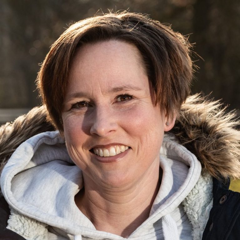 Kunde Tanja Frau mit lässiger Kleidung und Pelzkragen lächelt vor einer natürlichen Kulisse.