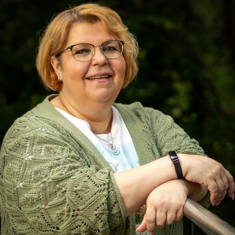 Kunde Silke Lächelnde Frau mit blonden Haaren, lässig gekleidet, steht an einem Geländer im Freien.