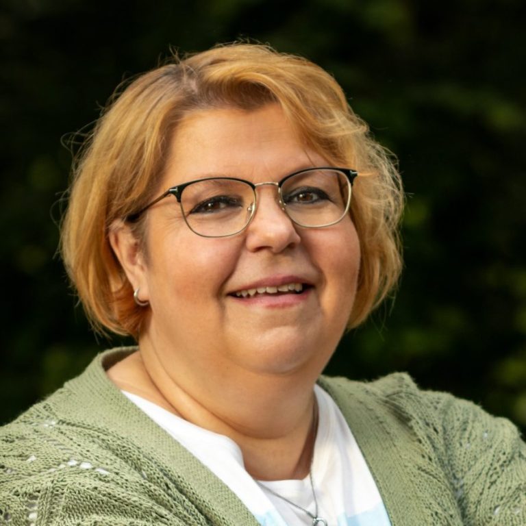 Kunde Silke Lächelnde Frau mit blonden Haaren, lässig gekleidet, steht an einem Geländer im Freien.
